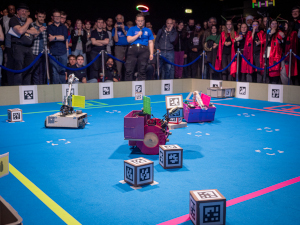 
            The SR2024 Final, viewed across the arena from the corner between Planets (scoring zones) 2 & 3.
            The venue lighting strongly illuminates the arena, which is surrounded on two sides by competitors and their supervisors.
            A volunteer commentator in a blue shirt, wearing a multicoloured LED halo, stands between the crowd and the arena.
            The edges of the arena are constructed from wooden panels, while the floor is blue carpet.
            The boundaries to the Planets are marked with brightly coloured tape.
            In the center of the frame a pink robot with a green flag for Planet 0 and large wheels holds up an Asteroid (cube shaped token).
            Just visible behind is the Egg (another token) on its Nest (a low plinth).
            To the left a robot with a yellow flag for Planet 3 and a tall arm heads away across the arena.
            To the right a pink robot with a pink flag for Planet 2 heads away across the arena.
            Scattered throughout the arena are other Asteroids -- some still on their starting positions and others moved by robots, either by accident or moved onto Planets.
            In the background, a Spaceship (a plastic box large enough for several Asteroids) is on Planet 1.
           